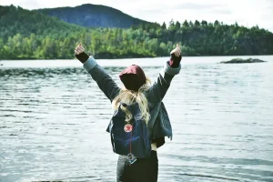 Mujer joven con los brazos en alto en señal de libertad frente a un lago con el fondo de un bosque