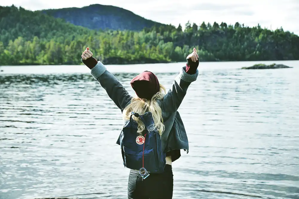 Mujer joven con los brazos en alto en señal de libertad frente a un lago con el fondo de un bosque