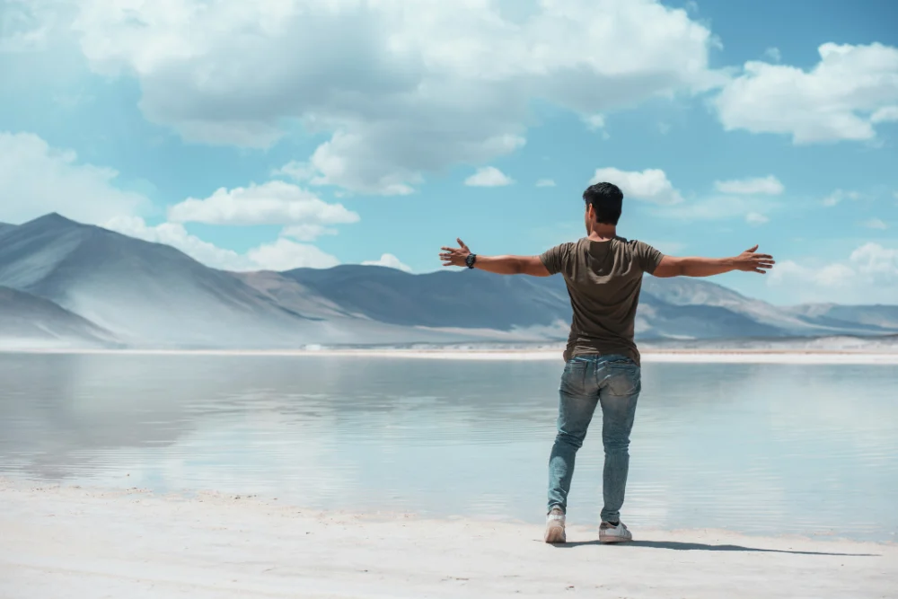 Hombre con los brazos abiertos en señal de libertad, dando la espalda a la cámara en un paisaje de un lago con el cielo celeste y un lago