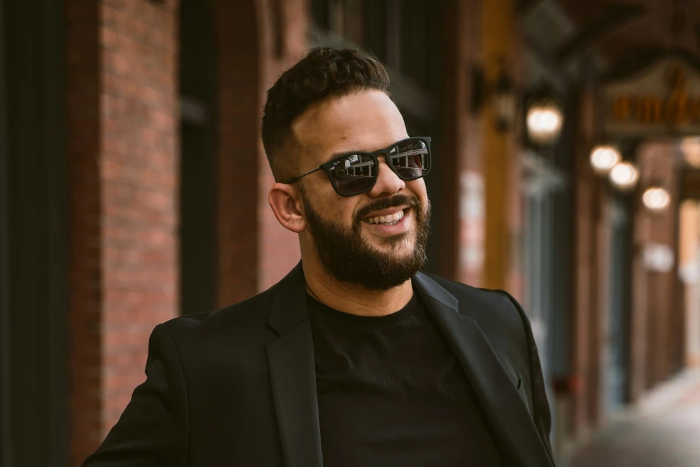 Hombre con vestido casual elegante con barba bien cuidada lentes oscuros y sonriendo.