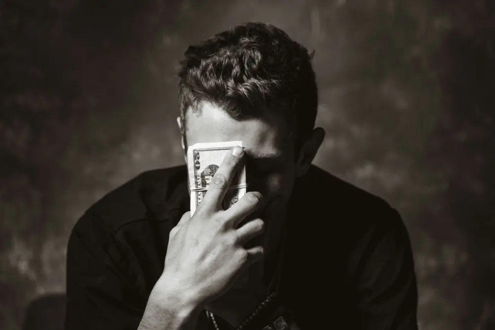 Hombre sentado mirando a la cara con un fajo de billetes de 100 dolares cubriendo su cara haciendo alución a impuestos en la inversión inmobiliaria